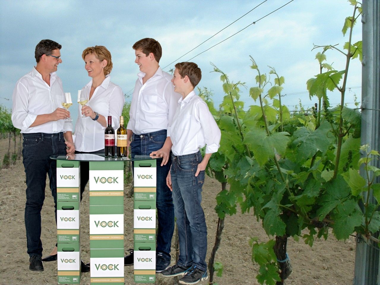 Four people are standing in a vineyard, clinking glasses of wine. In front of them is a table made from Vock wine cartons.