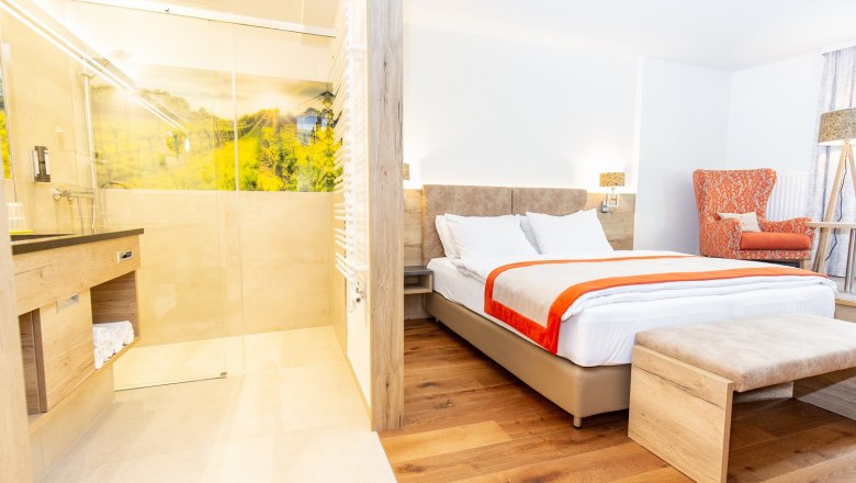 Modern hotel room with double bed, orange armchair and open shower.