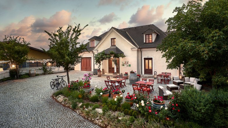 A cozy courtyard with tables, chairs and flower beds in front of a large house at sunset.
