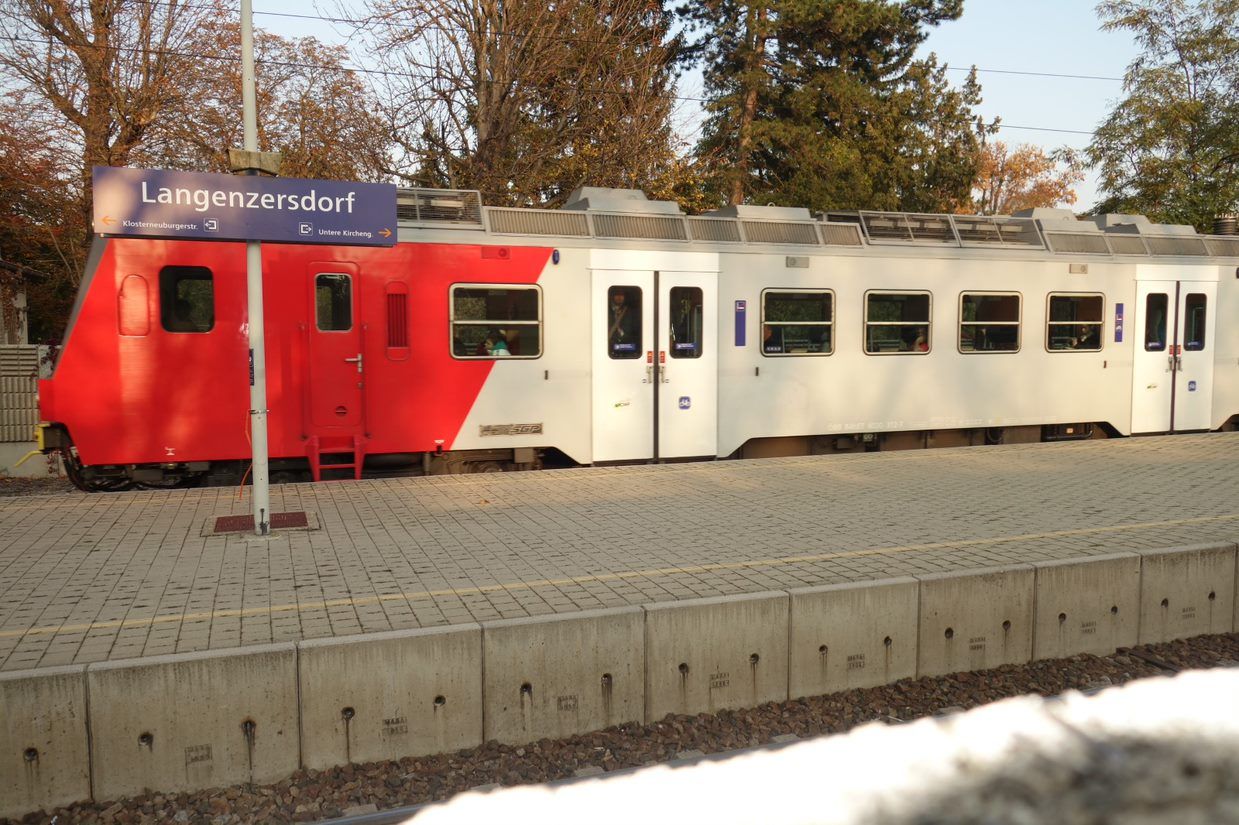 A train stands at Langenzersdorf station, with a sign in the foreground. The train is red and white and the surrounding area is surrounded by trees.