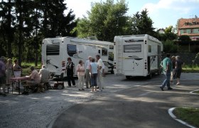 Menschen stehen und sitzen neben zwei Wohnmobilen auf einem Stellplatz im Freien.