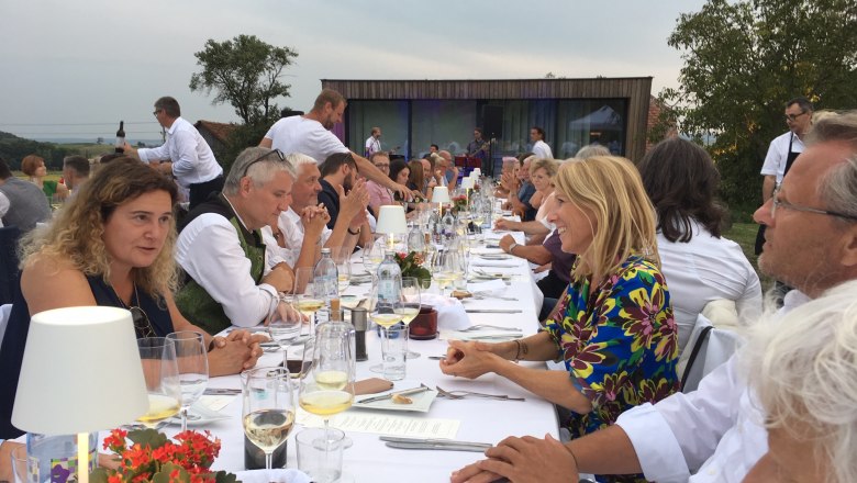 Menschen sitzen an einem langen Tisch im Freien und unterhalten sich bei einem festlichen Essen.