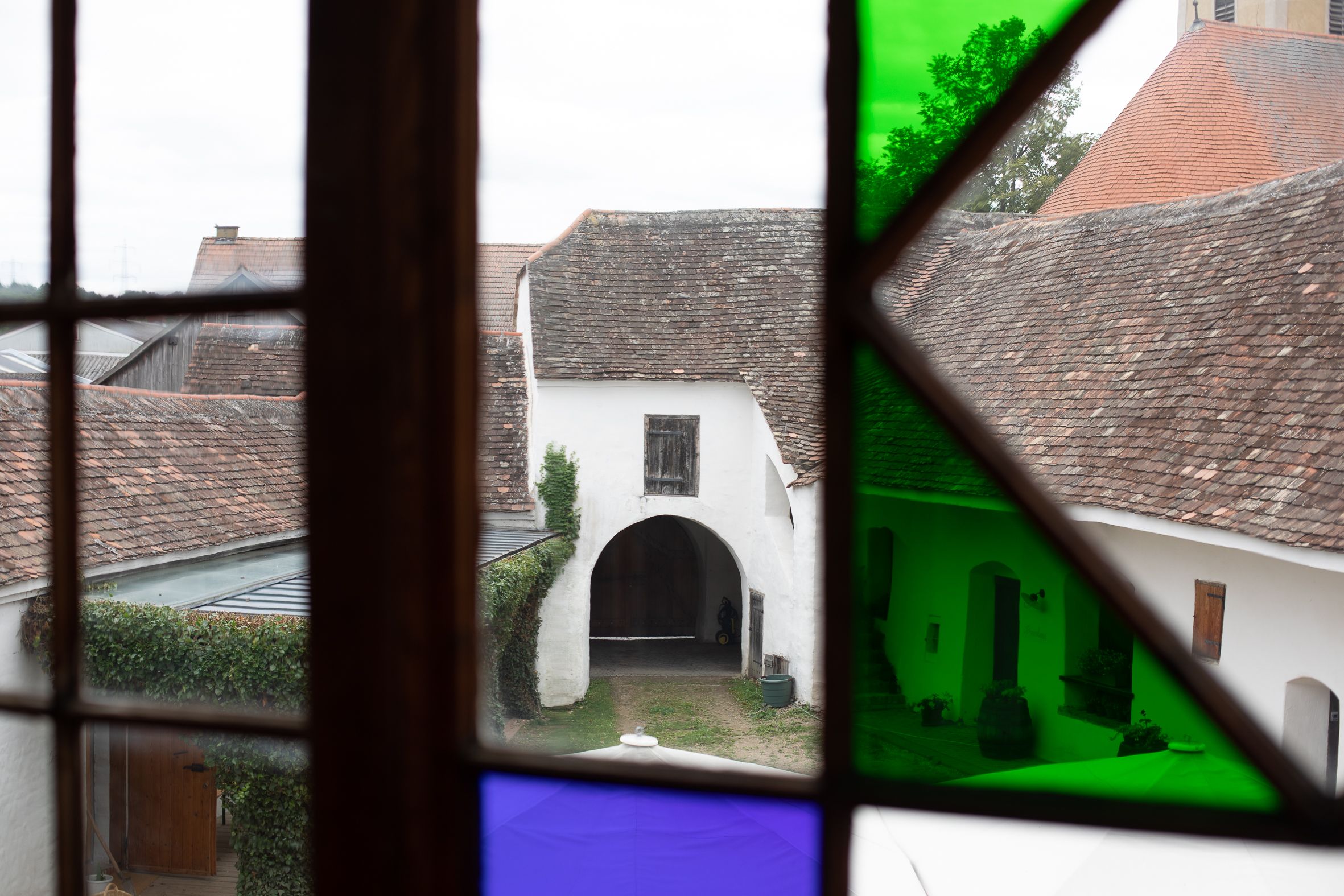 View through a stained glass window of an old courtyard with an arched gate and tiled roofs.