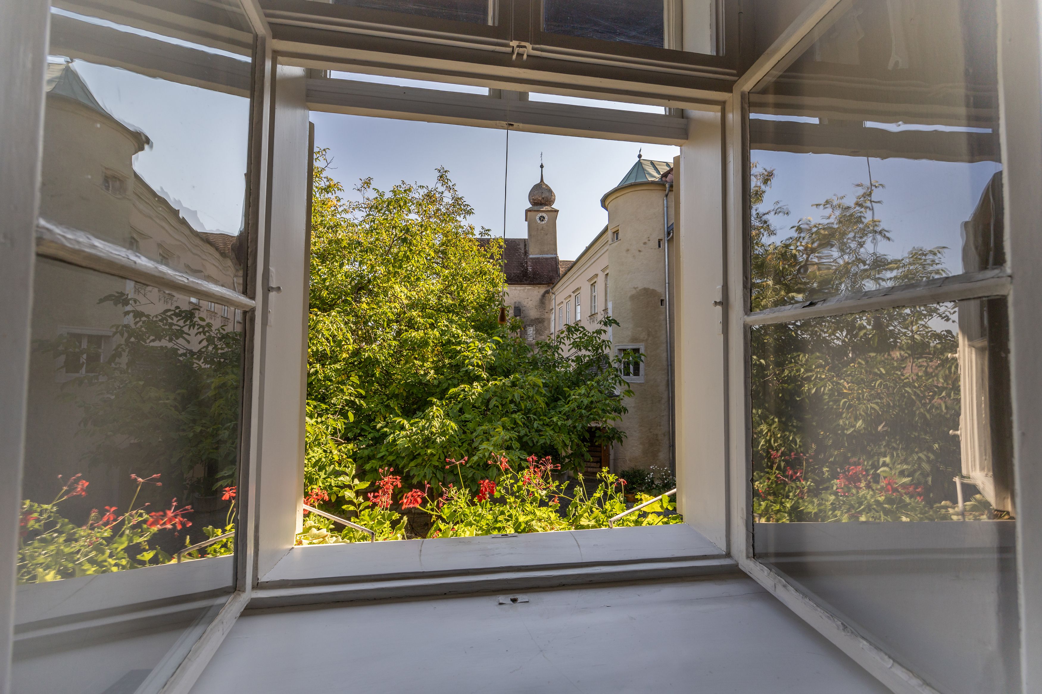 Blick aus einem offenen Fenster auf einen grünen Garten und ein historisches Gebäude mit Turm.