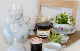 Frühstückstisch mit Teekanne, Honigglas, Marmelade, Eiern und Blumen.