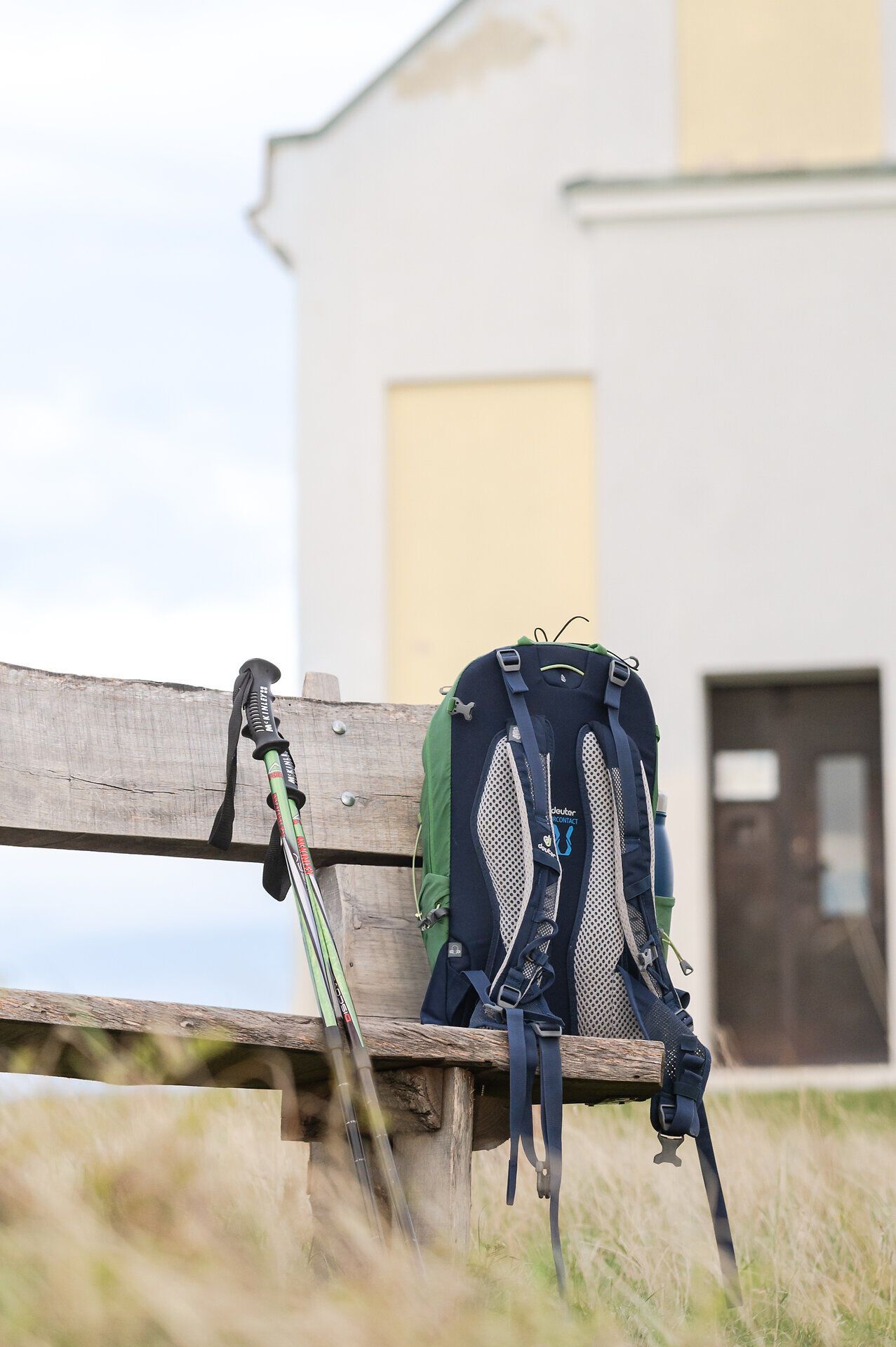 Ein Wanderrucksack steht auf einer Holzbank, daneben lehnen Wanderstöcke, während im Hintergrund der Anschnitt einer Kapelle erkennbar ist.