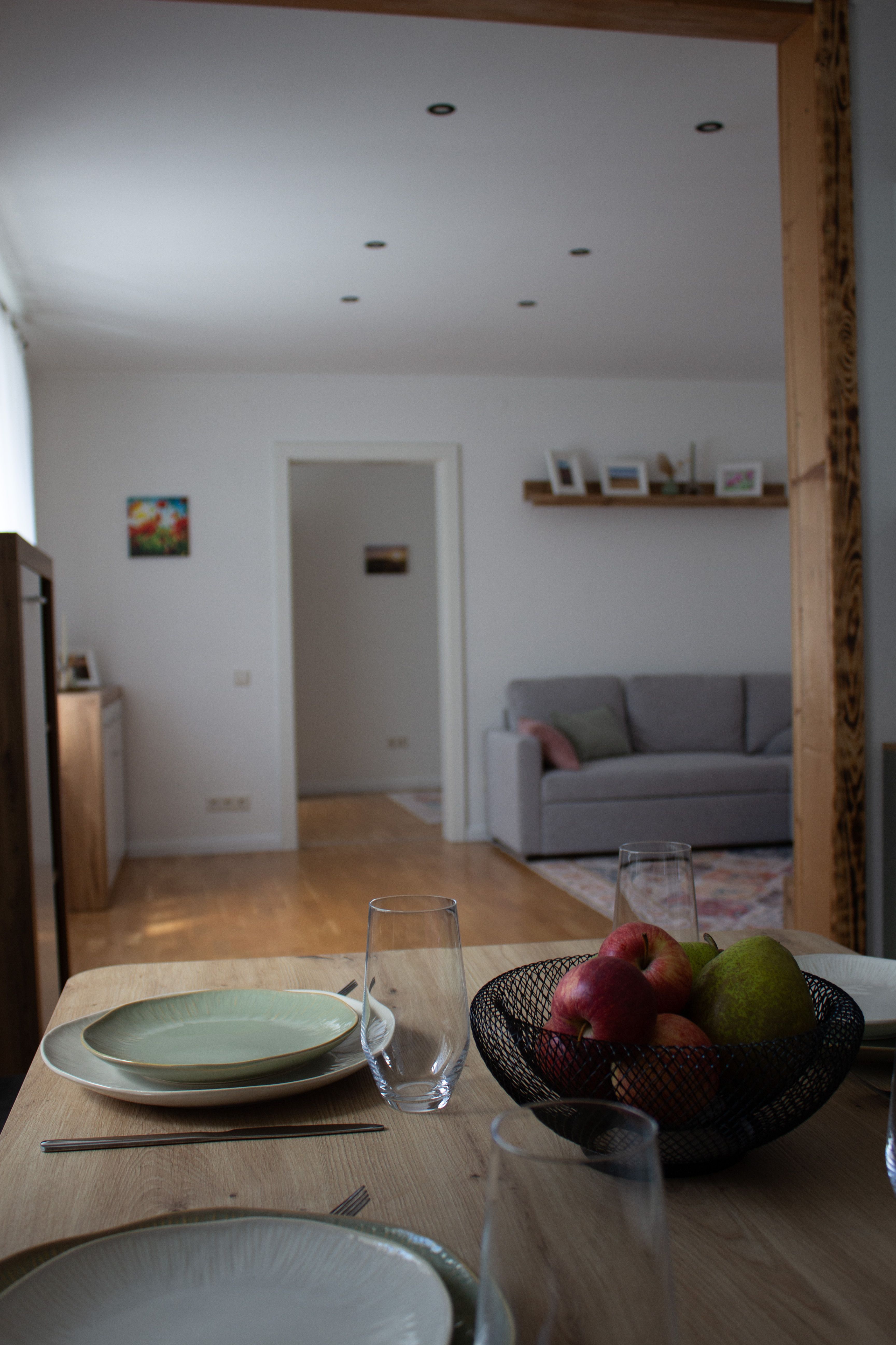 A dining area with set table, fruit bowl and view of a living room with gray sofa.