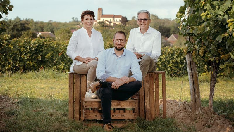 Drei Personen sitzen auf Holzkisten in einem Weingarten, im Hintergrund eine Kirche.