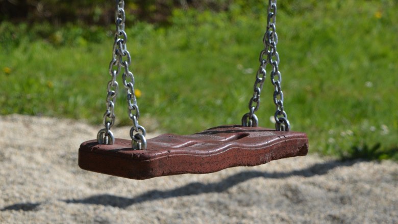 Nahaufnahme einer leeren Schaukel aus Holz mit Metallketten, die über einem Kiesboden hängt. Im Hintergrund ist unscharfes Gras zu sehen.