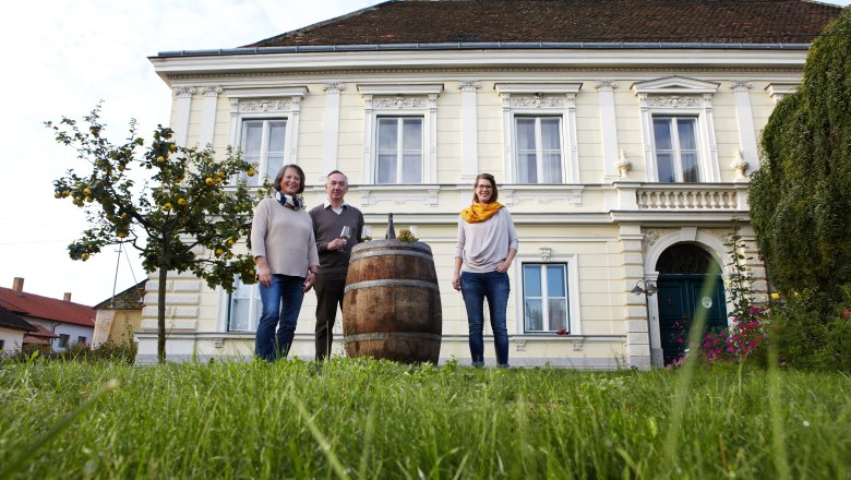 Drei Personen stehen vor einem großen, gelben Gebäude mit einem Fass im Vordergrund.