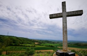 Ein großes Steinkreuz auf einem Hügel mit Blick auf eine grüne Landschaft und bewölkten Himmel.