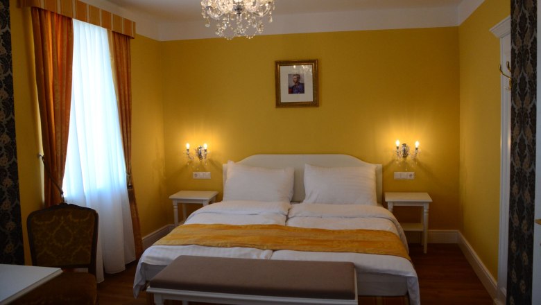 Elegantes Hotelzimmer mit gelben Wänden, Doppelbett, Kronleuchter und Bild an der Wand.