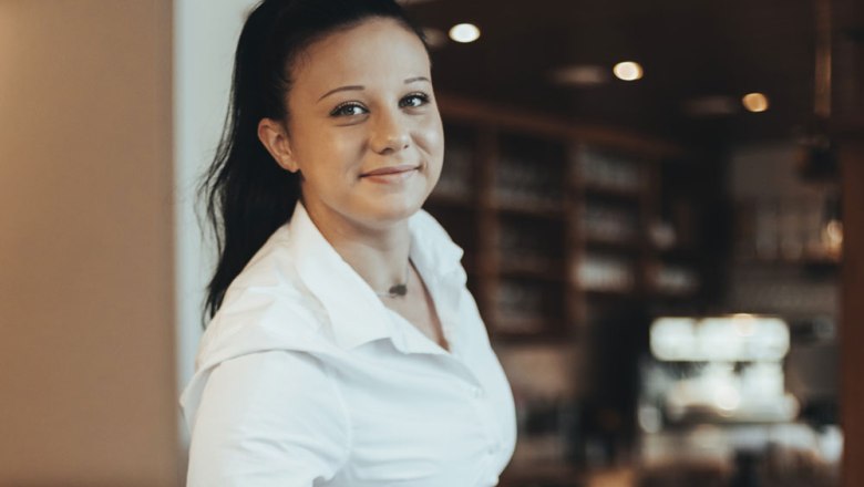 Frau in weißem Hemd lächelt in die Kamera, Hintergrund unscharf.