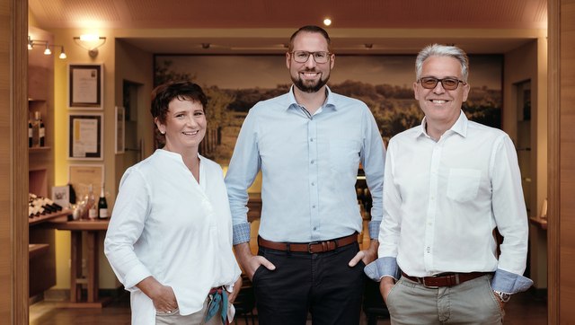 Drei Personen in einem Raum mit Weinflaschen im Hintergrund.