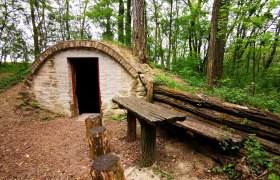 Steinhütte im Wald mit Holztisch und Baumstümpfen als Sitzgelegenheiten.