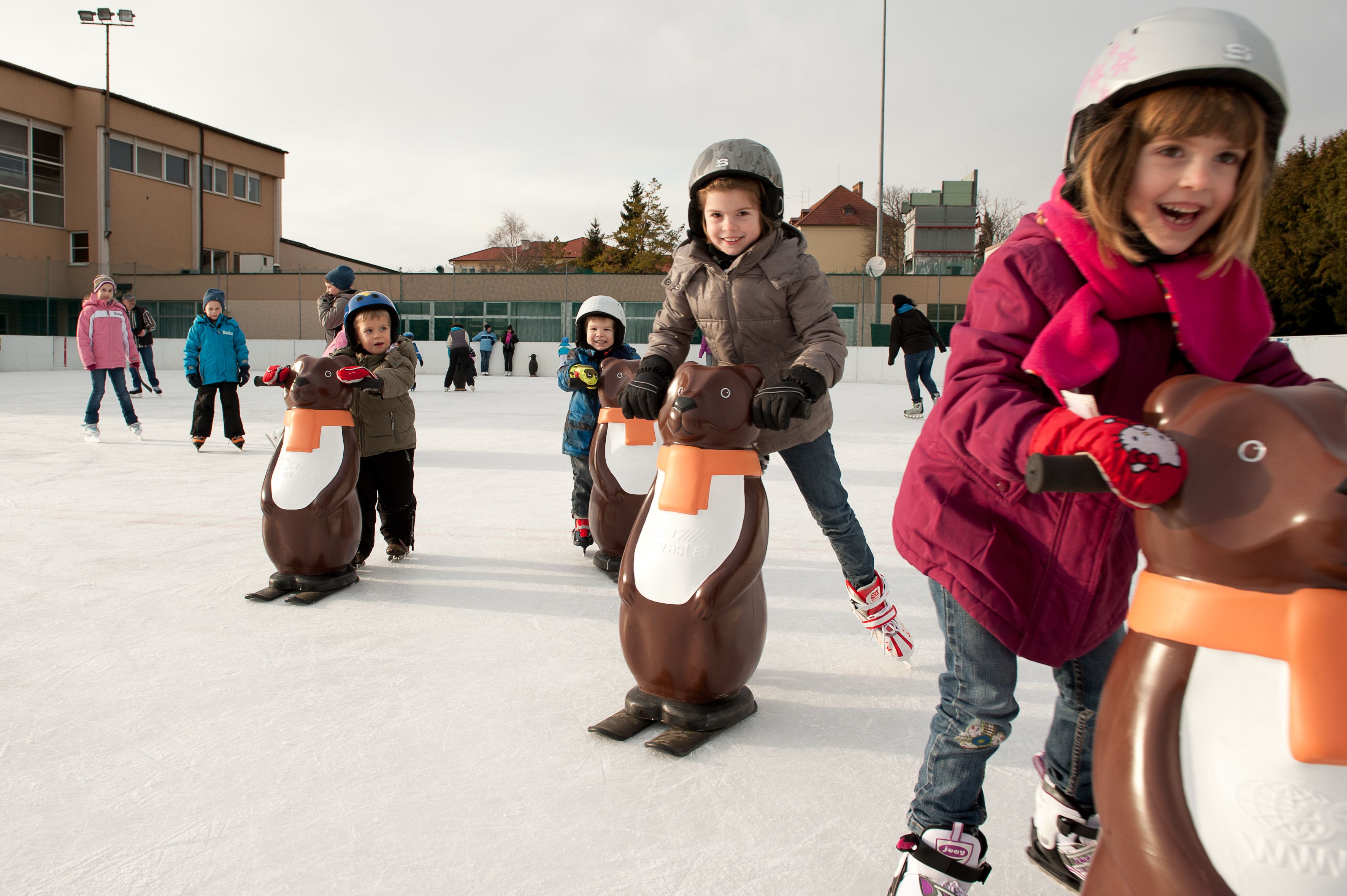 Kinder beim Schlittschuhlaufen mit Pinguin-Hilfen auf einer Eisbahn.
