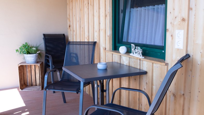 Ein Balkon mit Holzw&auml;nden, zwei schwarzen St&uuml;hlen, einem Tisch und einer Pflanze in einem Topf.