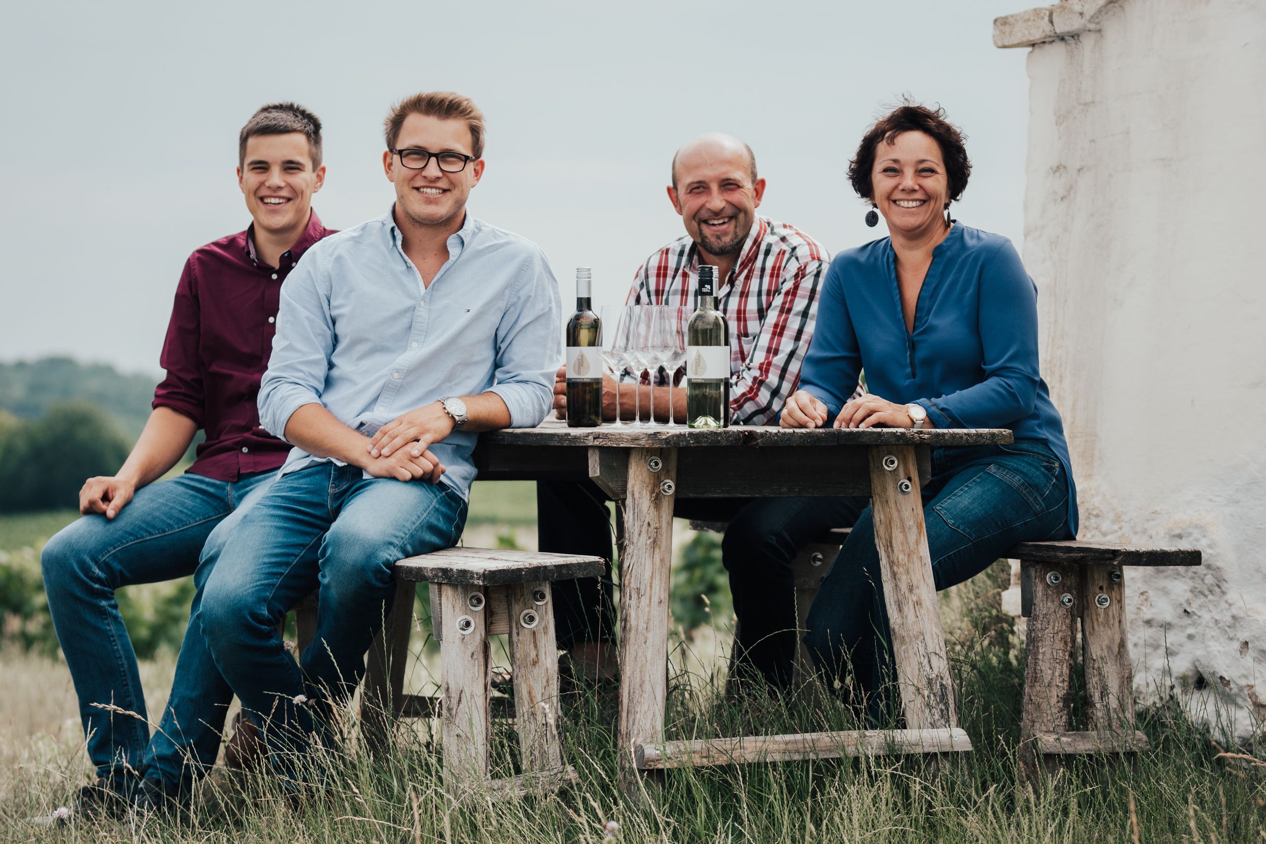 Four people sit at a wooden table outside with bottles of wine and glasses.