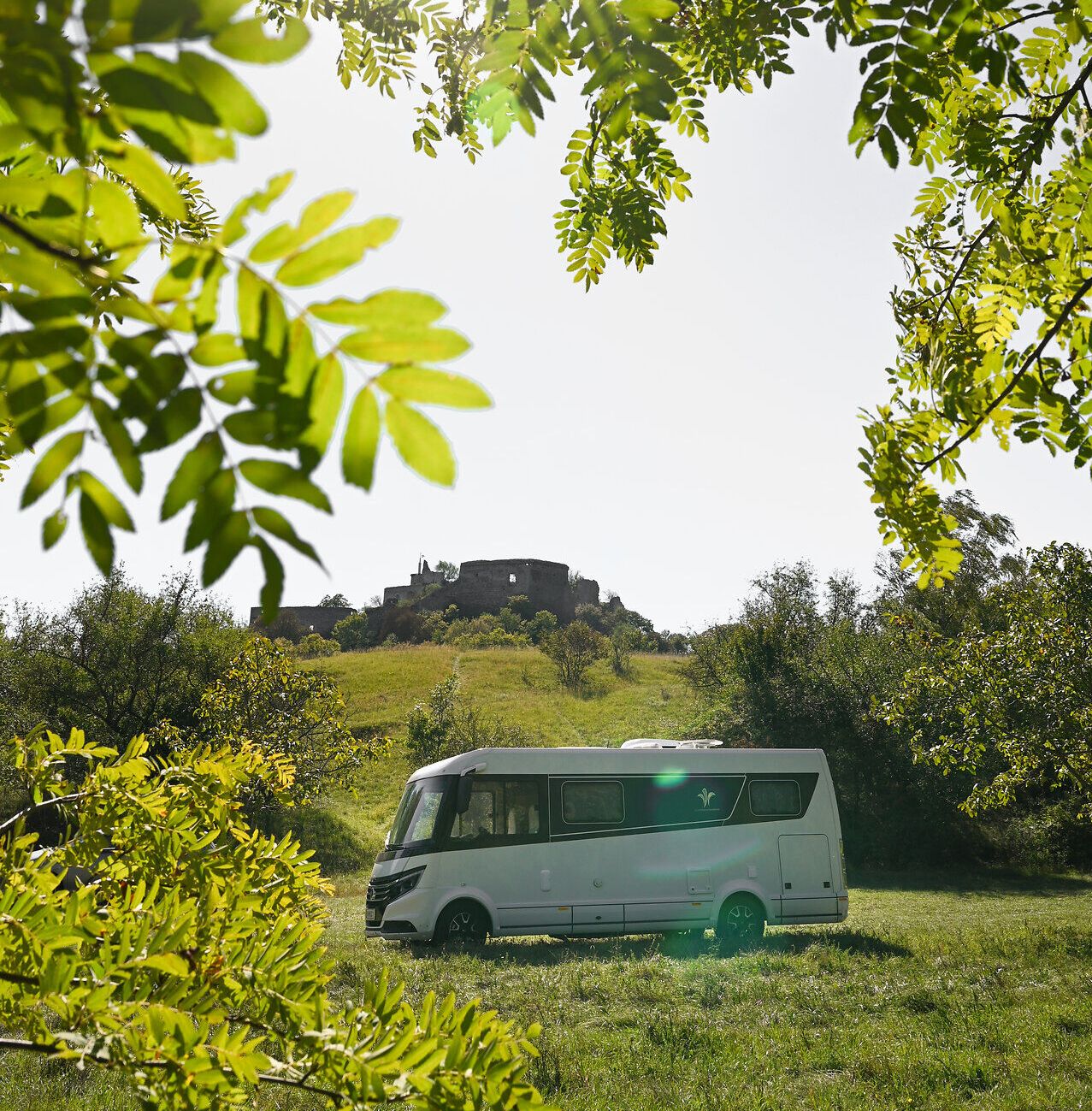 Umgeben von üppigem Grün und unter einem strahlend blauen Himmel, bietet der Campingwagen einen perfekten Rückzugsort für Naturliebhaber. Die sanften Hügel im Hintergrund und die historische Burgruine schaffen eine malerische Kulisse, die zum Entspannen und Erkunden einlädt.