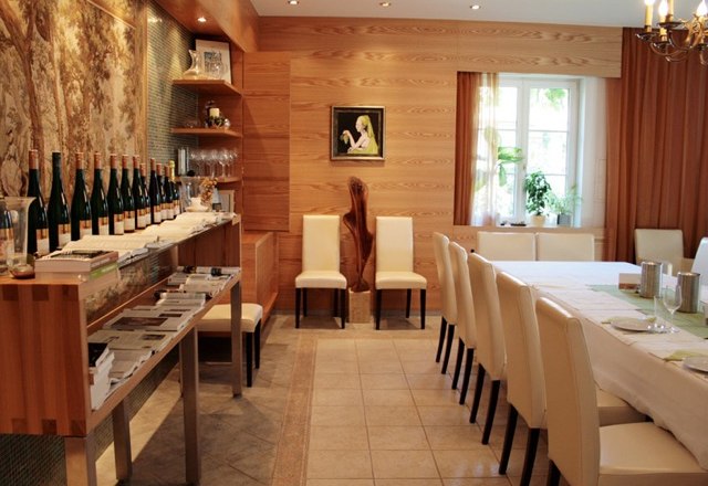 Ein Verkostungsraum mit Holzwänden, einem langen Tisch mit weißen Stühlen und Weinflaschen auf einem Regal.