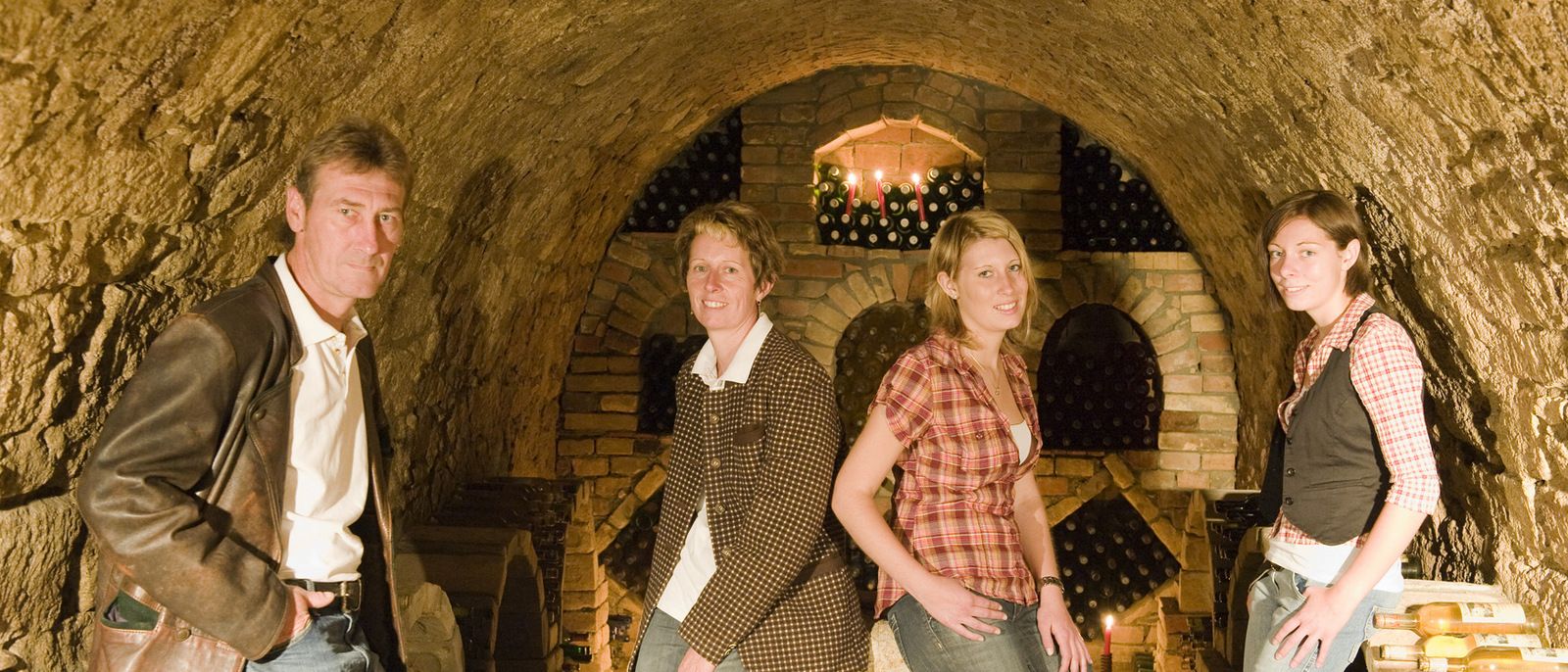 Four people in a wine cellar with brick walls and wine bottles in the background.