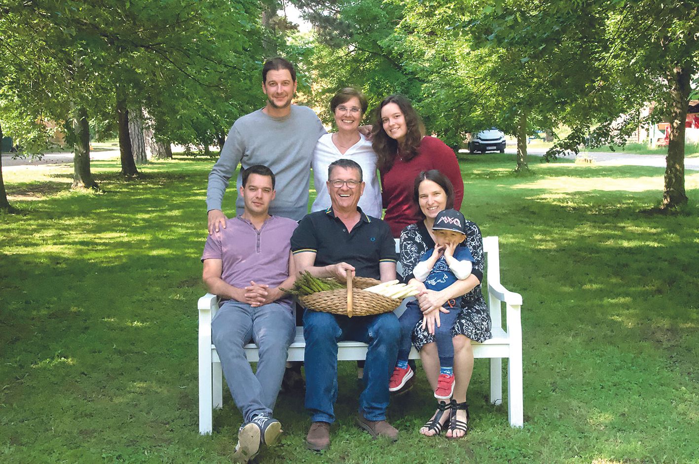 A family is sitting on a bench in the countryside, surrounded by trees. A man holds a basket of asparagus. A child sits on a woman's lap.