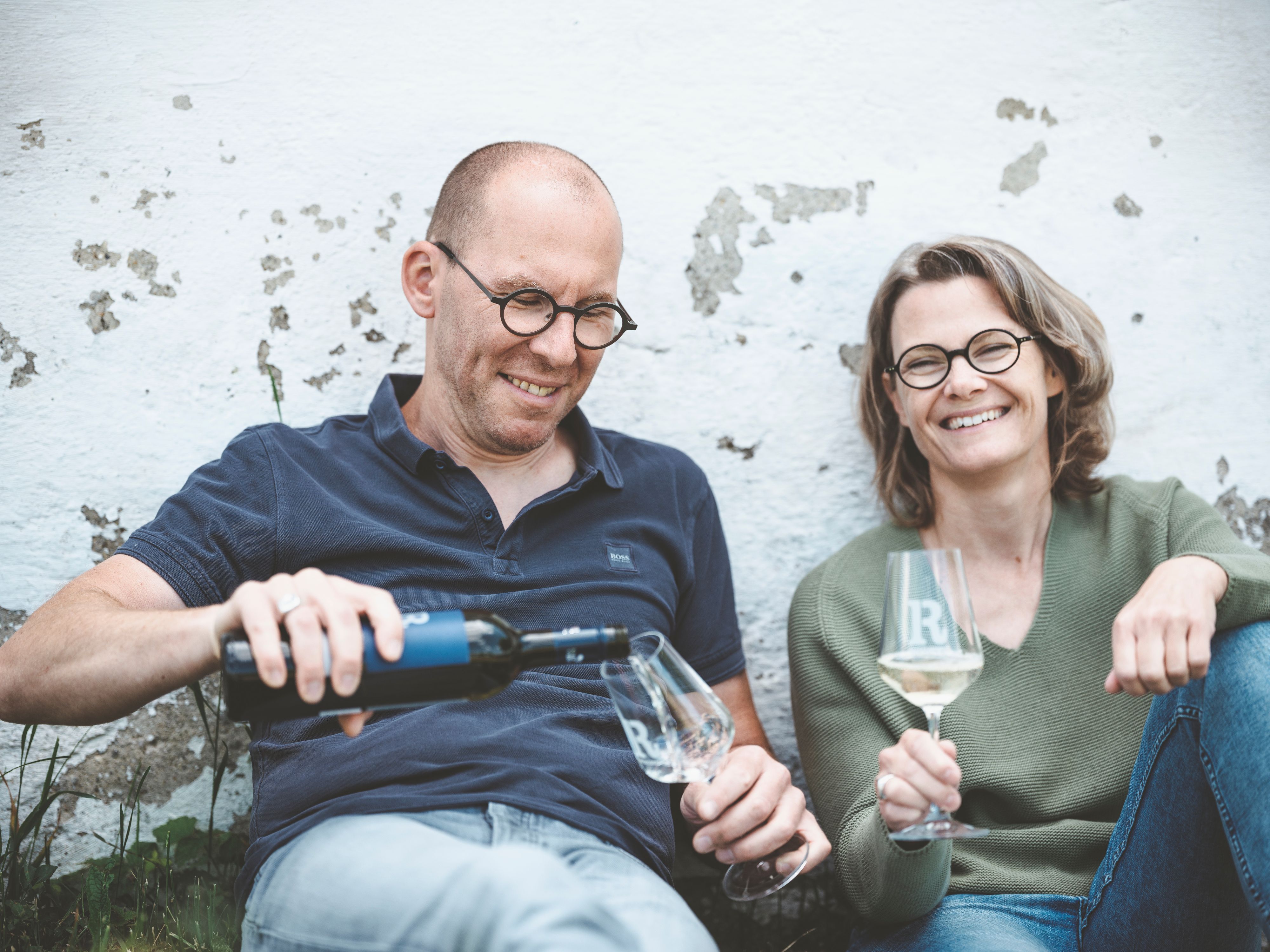 Two people sit relaxed against a wall and drink wine.