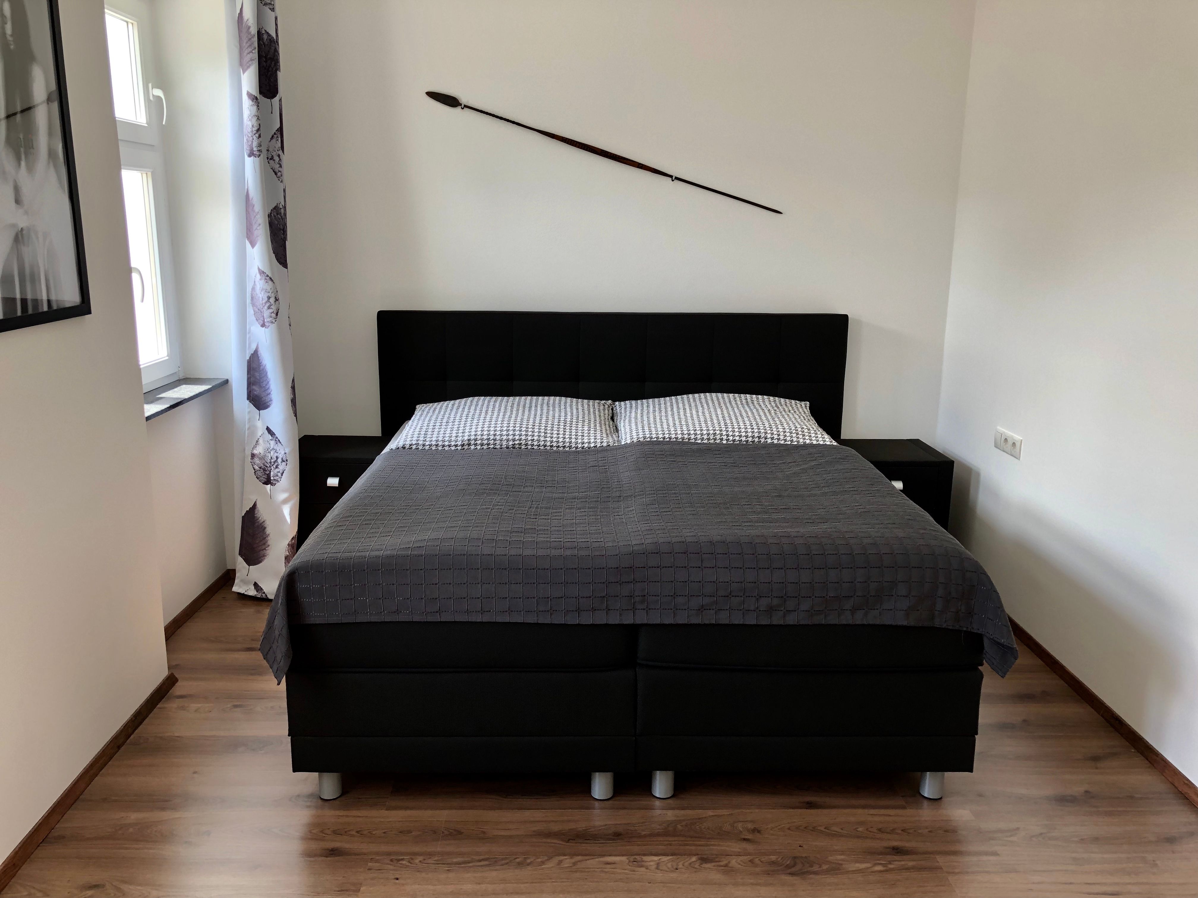 Modernes Schlafzimmer mit einem großen Bett, grauer Bettdecke und einem Speer an der Wand.