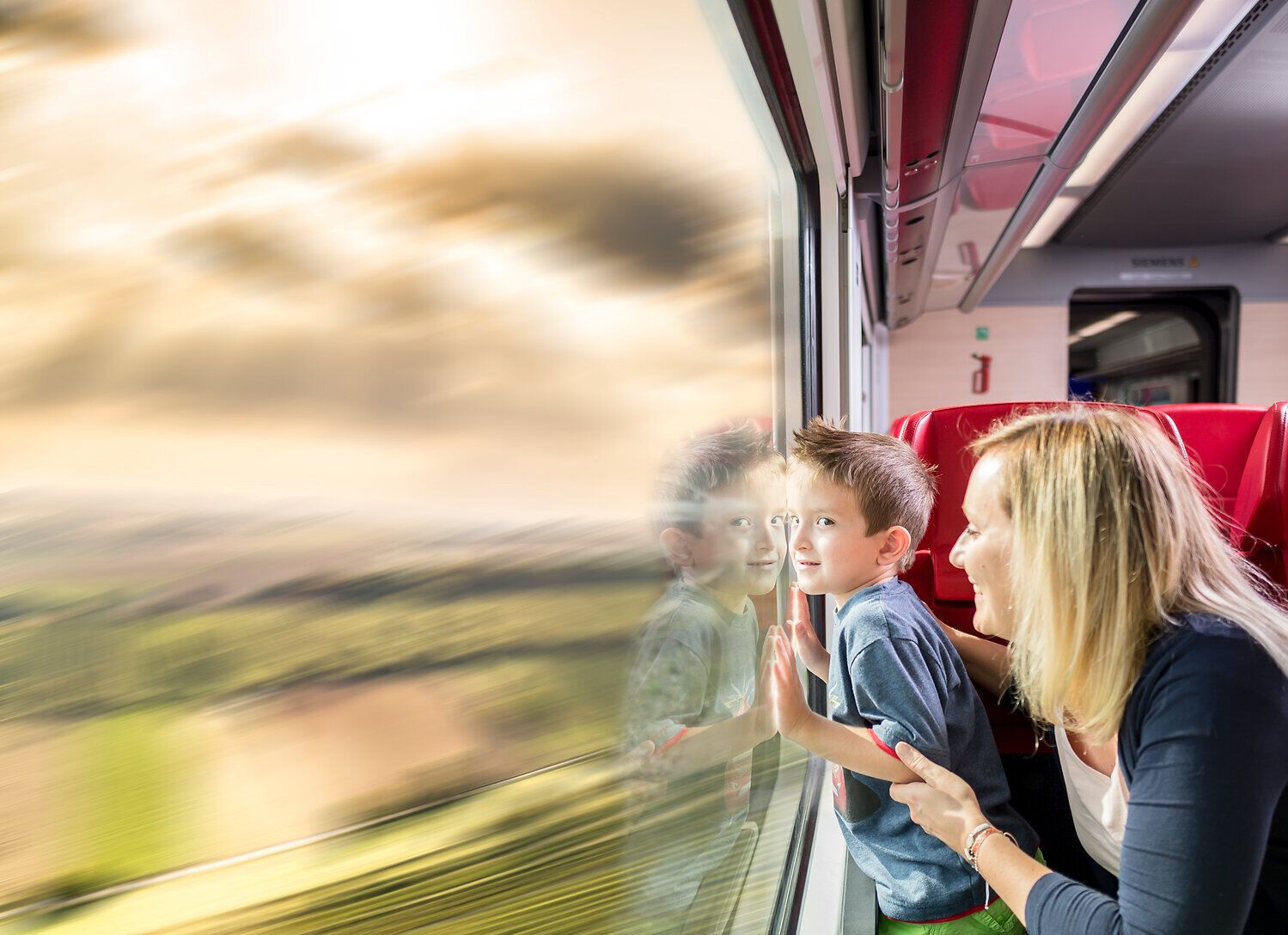 Eine Familie genießt die malerische Aussicht während ihrer Bahnfahrt. Der Junge drückt seine Nase an das Fenster, während die grünen Wiesen und sanften Hügel vorbeiziehen. Ein perfekter Moment, um die Schönheit der Natur zu erleben und gemeinsame Erinnerungen zu schaffen.