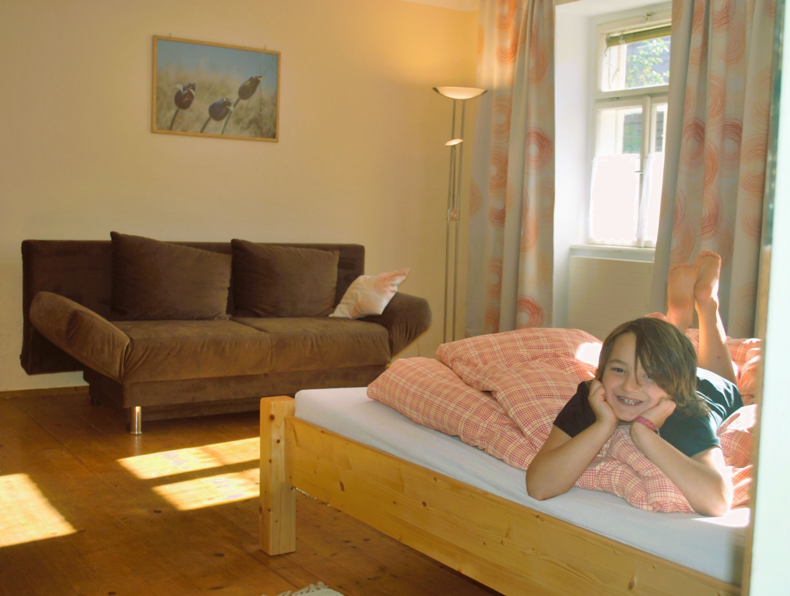 Ein Kind liegt lächelnd auf einem Bett in einem gemütlichen Zimmer mit Sofa und Bild an der Wand.
