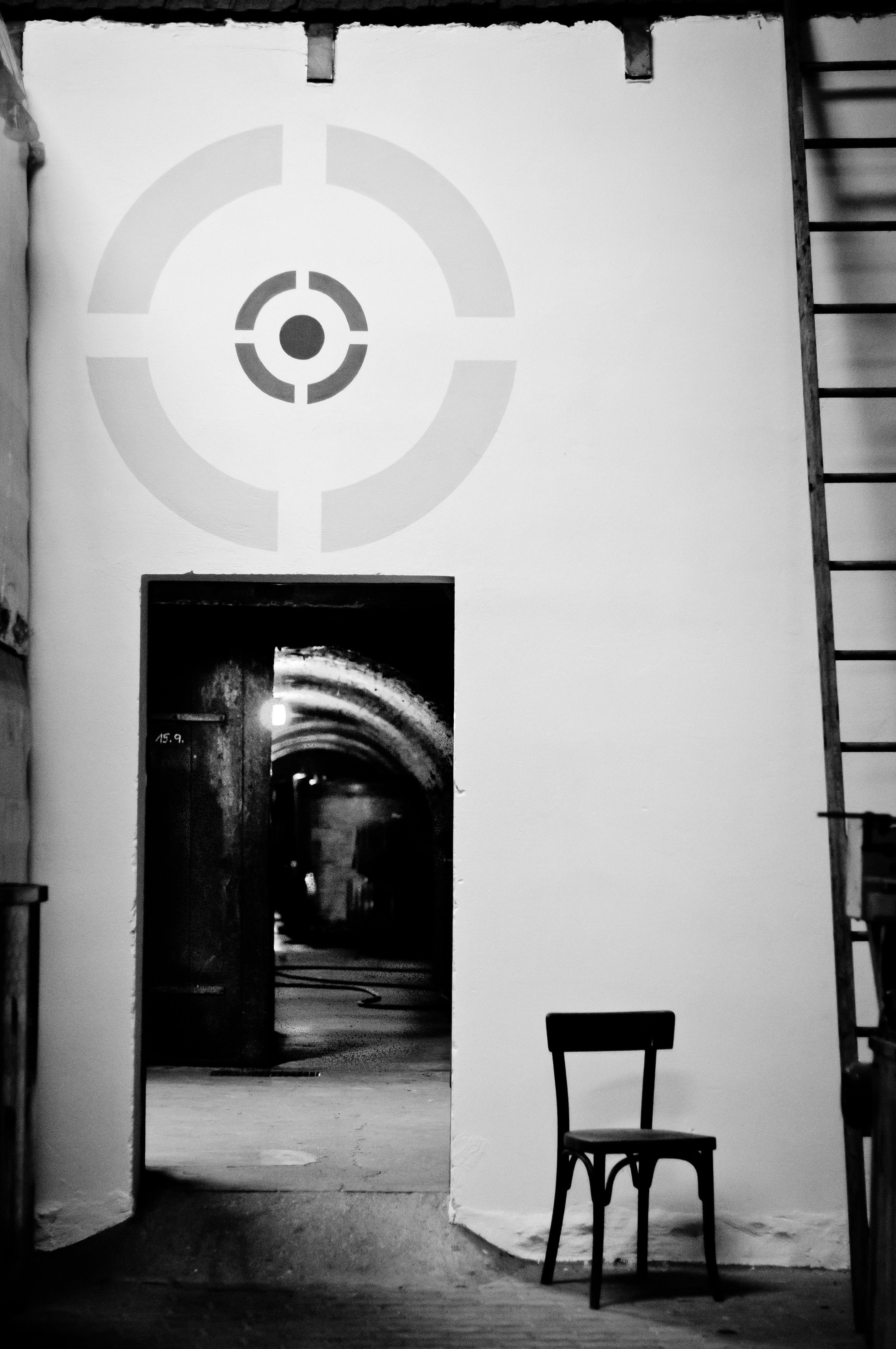 A black and white photo of a cellar with a chair in front of a door and a target pattern on the wall.