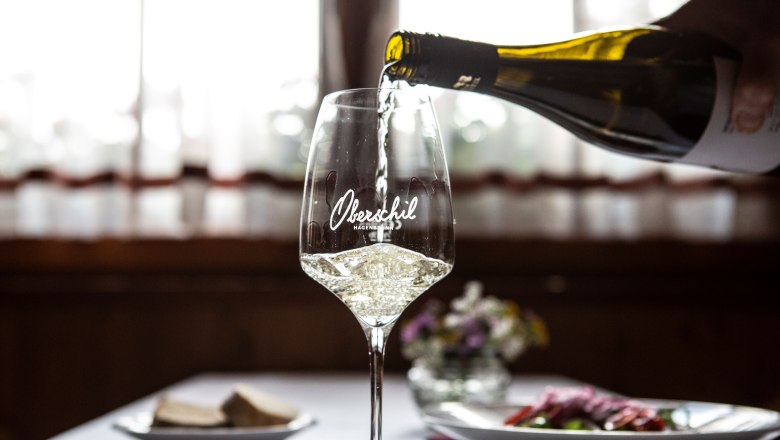 A glass of white wine is poured, a table laid with flowers in the background.