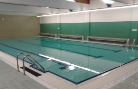 Innenansicht eines leeren Hallenbads mit grün gefliesten Wänden und einem großen Schwimmbecken.