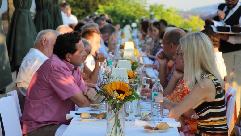 Menschen sitzen an einer langen, festlich gedeckten Tafel im Freien.