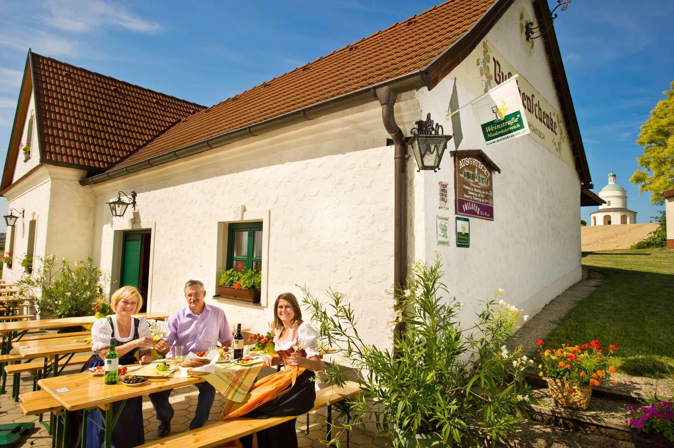 Drei Personen sitzen an einem Tisch vor einem Weingut und genießen eine Mahlzeit im Freien.