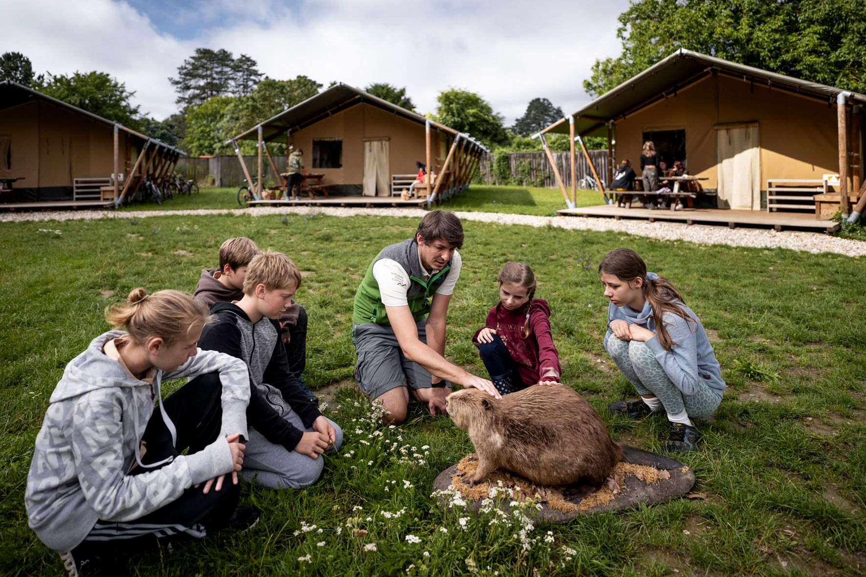 Eine Gruppe von Kindern und ein Erwachsener sitzen auf einer Wiese und streicheln ein großes Nagetier. Im Hintergrund sind Zelte zu sehen.