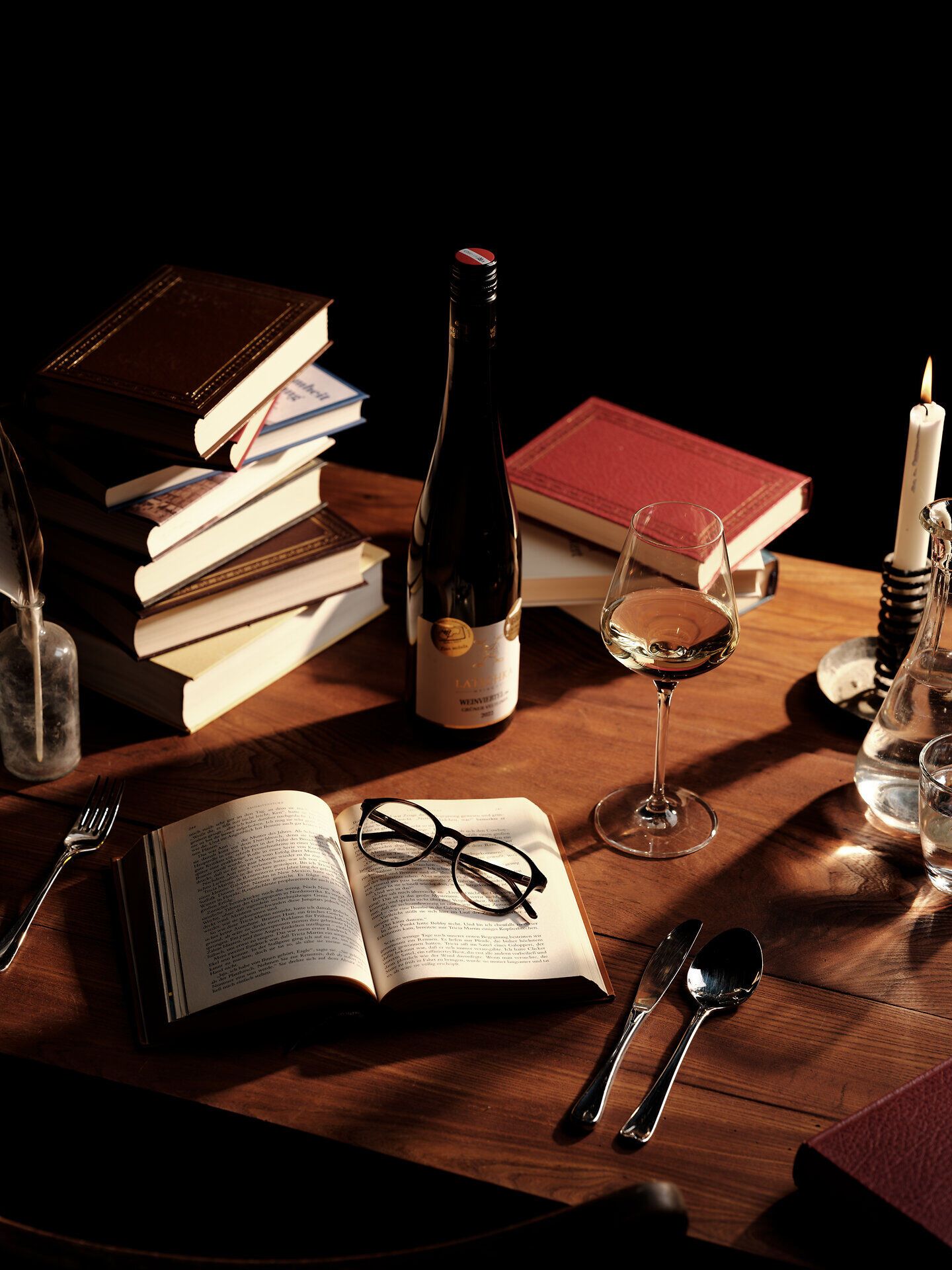 Ein einladendes Stillleben, das die Gemütlichkeit des Herbstes verkörpert. Ein offenes Buch, umgeben von einer Flasche Wein und einem Glas, lädt dazu ein, in die Welt der Geschichten und Aromen einzutauchen. Kerzenlicht und die sanfte Präsenz von Büchern schaffen eine harmonische Atmosphäre, die zum Verweilen einlädt.
