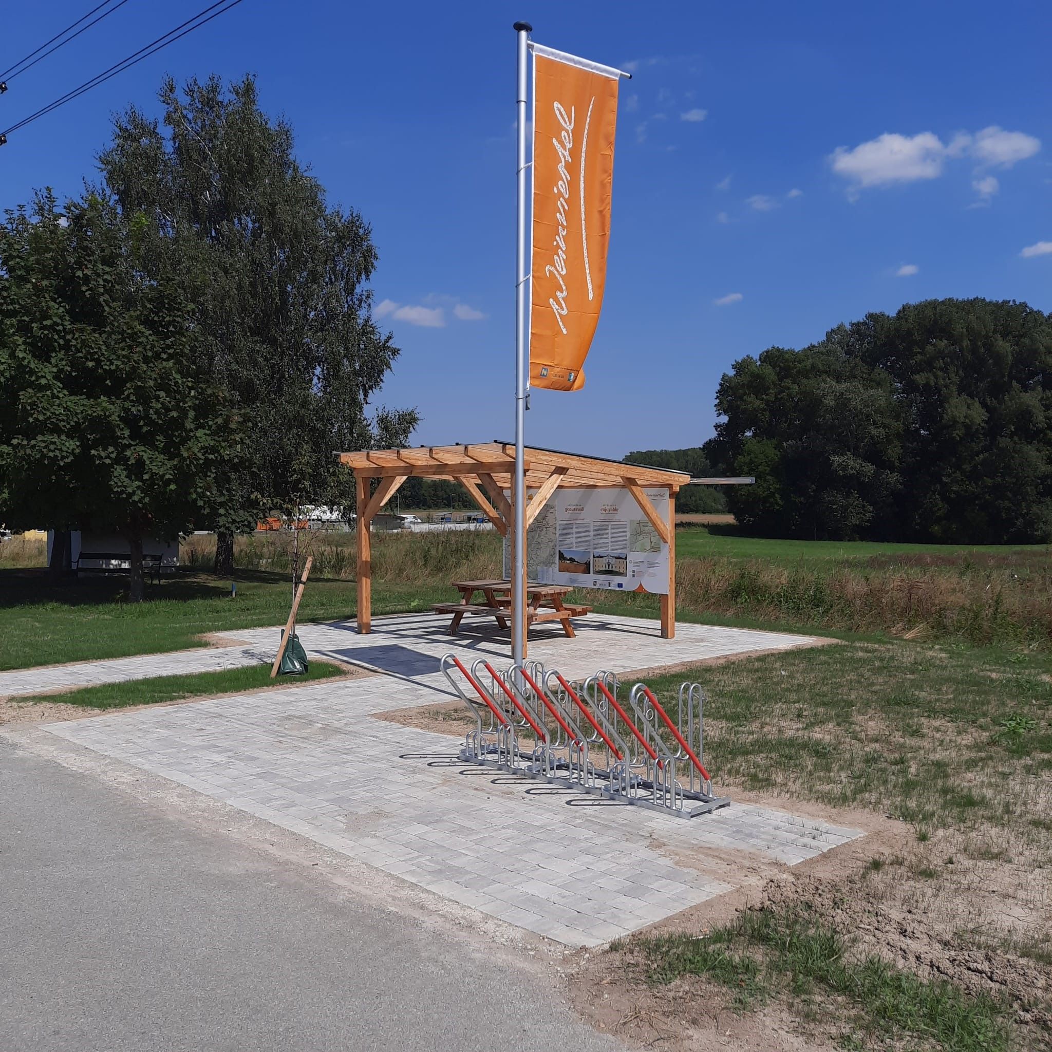 Ein Rastplatz mit einer Holzpergola, einem Tisch und Fahrradständern. Eine orange Fahne mit der Aufschrift 'Weinviertel' weht im Wind.