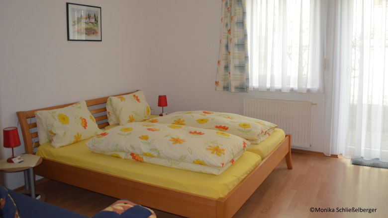 Ein gem&uuml;tliches Doppelzimmer mit einem Holzbett, bunter Bettw&auml;sche und zwei roten Nachttischlampen. Ein Fenster mit Vorh&auml;ngen ist im Hintergrund.