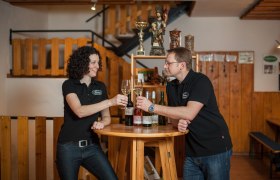 Zwei Personen sto&szlig;en mit Weingl&auml;sern an einem Holztisch an.