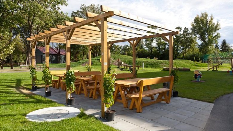Holzpergola mit Bänken und Tischen im Freien, umgeben von Rasen und Bäumen.