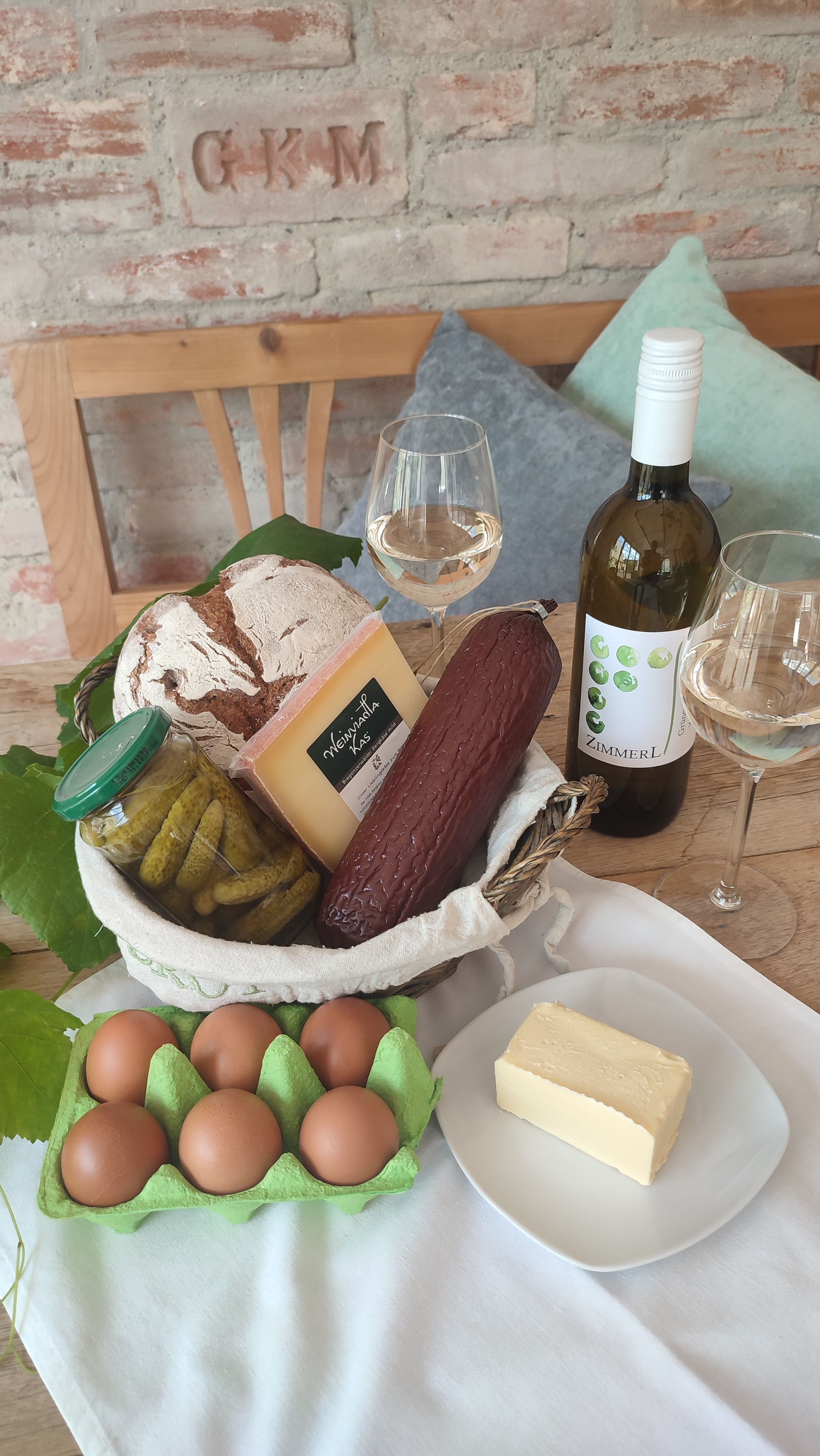 Ein Tisch mit regionalen Lebensmitteln: Brot, Käse, Wurst, Gurken, Eier, Butter und eine Flasche Weißwein mit zwei Gläsern.