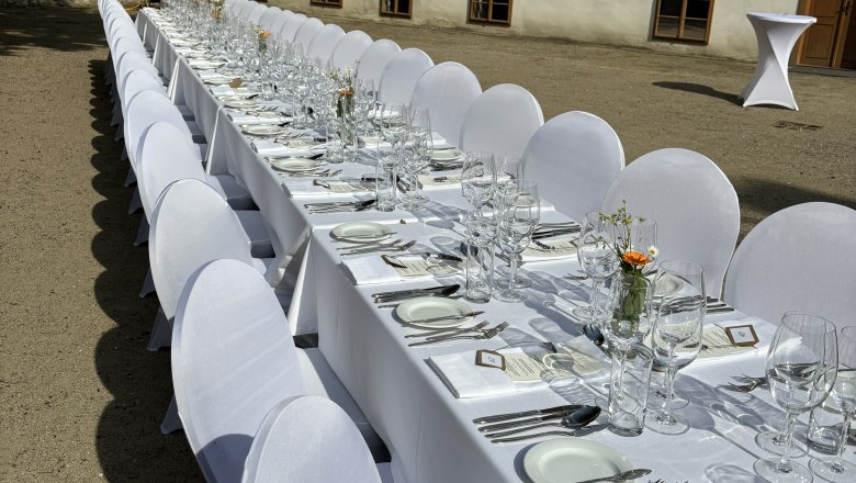 Lange Tafel im Freien mit weißer Tischdecke und Gedecken vor einem historischen Gebäude.