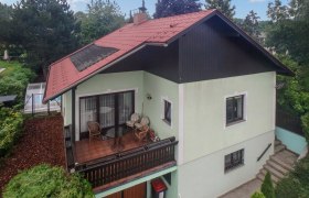 Zweistöckiges Haus mit rotem Dach und Balkon, umgeben von Bäumen.