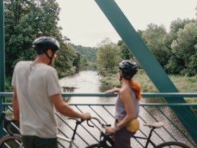 Zwei Radfahrer mit Helmen stehen auf einer Brücke und blicken auf einen Fluss, umgeben von üppiger Vegetation.
