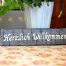 Ein Holzschild mit der Aufschrift 'Herzlich Willkommen' steht auf einem blumigen Tisch.