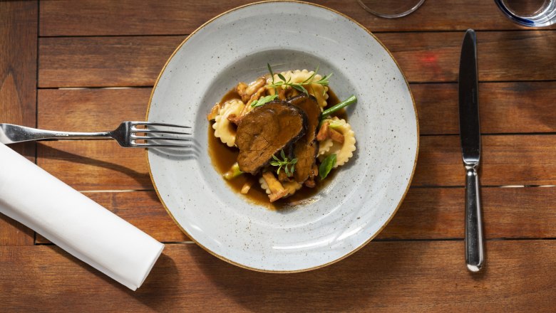 Ein Teller mit geschmorten Rindsbackerl, Ravioli und Gemüse auf einem Holztisch, daneben Besteck und ein Glas Rotwein.