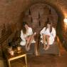Two women in white bathrobes relax on loungers in a wine cellar with brick walls.
