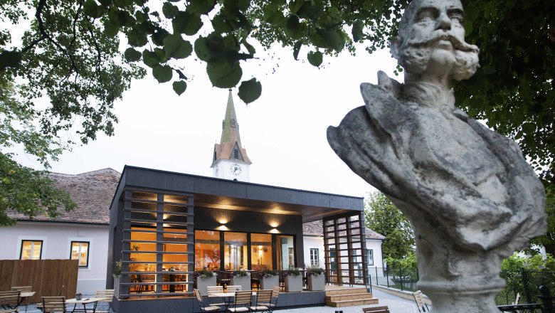 Büste vor modernem Gebäude mit Kirchturm im Hintergrund, umgeben von Bäumen.