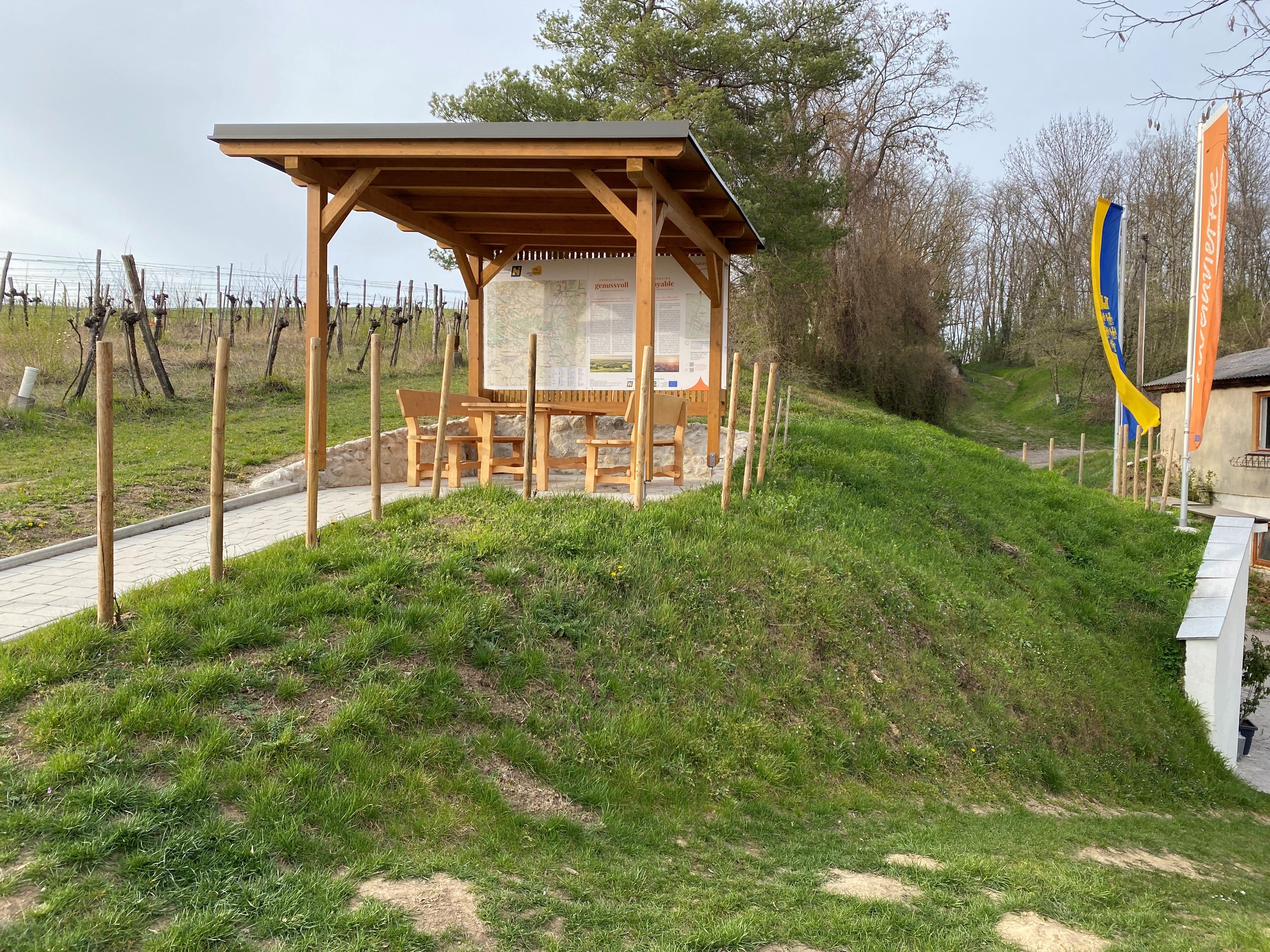 A rest area with a wooden roof and seating on a grassy hill, surrounded by vineyards and trees.